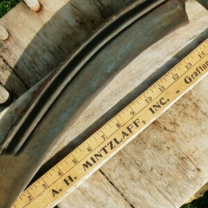May include: A vintage, curved metal blade with a wooden ruler. The blade is a dark, weathered grey with a slight curve. A wooden ruler with black markings and the text "A. H. MINTZLAFF, INC. - Grafton, Wis." is placed next to the blade.