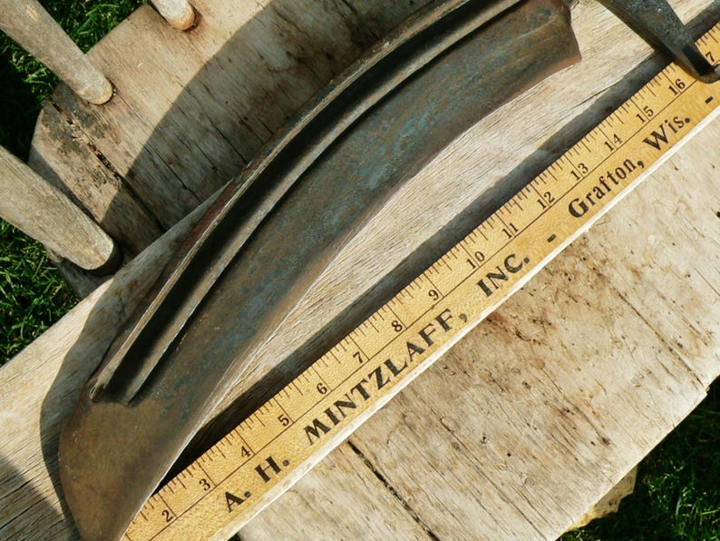 May include: A vintage, curved metal blade with a wooden ruler. The blade is a dark, weathered grey with a slight curve. A wooden ruler with black markings and the text "A. H. MINTZLAFF, INC. - Grafton, Wis." is placed next to the blade.