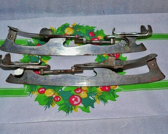 May include: Two vintage ice skate blades with a silver finish. The blades are attached to a metal frame with a screw and nut. The blades are on a white surface with a green and red floral design.