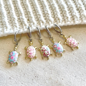 May include: Six gold turtle stitch markers with floral patterns in pink and blue. The markers are hanging from a white crocheted fabric.