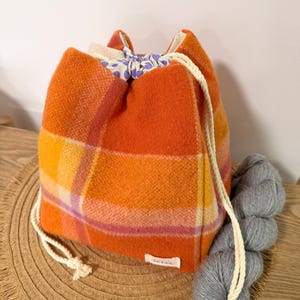 May include: A drawstring bag made of orange, yellow, and pink plaid wool fabric. The bag features a white rope closure and a light-coloured interior lining with a purple heart pattern. A small white rectangular label is attached to the bottom corner.