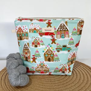 May include: A light blue zipped pouch featuring a repeating pattern of gingerbread houses, gingerbread people, Christmas trees, and vintage trucks. A small rectangular fabric label with the text made by kate & adele is centred on the front.