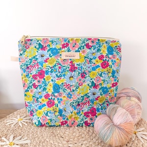 May include: A zipped pouch featuring a vibrant floral pattern with pink, yellow, and blue flowers on a light background. A small rectangular fabric label with the text "kate adela" is centred on the front. A gold-toned metal zip is at the top.