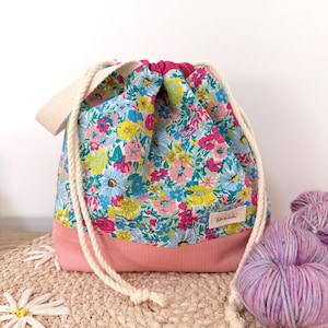 May include: A drawstring bag featuring a vibrant floral pattern with blue, pink, and yellow flowers on the upper section and a solid dusty pink base. The bag includes thick cream-coloured rope drawstrings and a small rectangular fabric label.