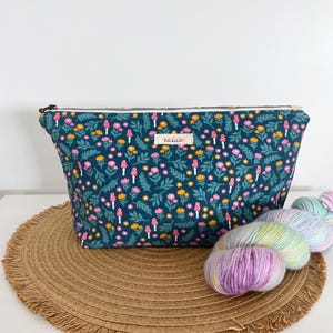 May include: A teal floral print pouch with a zipper closure, featuring a pattern of pink, yellow, and white flowers and mushrooms. A skein of colorful yarn rests beside the pouch on a woven mat. The pouch has a small label that reads "tete-a-tete".