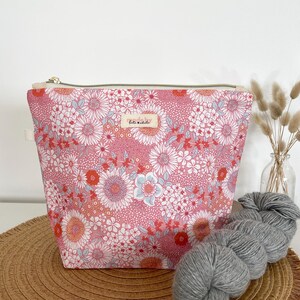 May include: A pink floral print zippered pouch with a light-coloured interior. The pouch is next to a skein of grey yarn. The pouch has a small rectangular label with text. The background includes a woven mat and a vase with dried flowers.