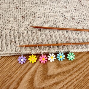 May include: Six metal stitch markers featuring small, colourful daisy-shaped charms in purple, yellow, pink, white, light blue, and mint green. The markers are attached to two wooden knitting needles resting against a textured beige knit fabric.