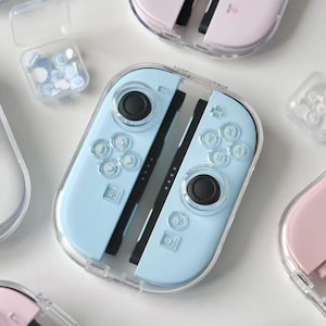 Puede incluir: Una variedad de mandos Joy-Con de Nintendo Switch en colores pastel dentro de fundas protectoras transparentes. Los mandos son de color rosa, azul y blanco, con joysticks y botones negros. También se ven pequeños recipientes con agarres para pulgar a juego.