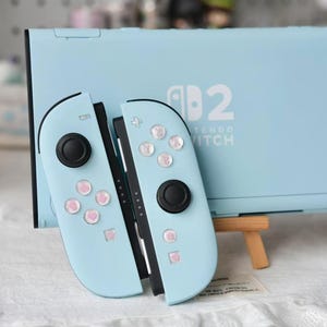 May include: A light blue Nintendo Switch console and two matching Joy-Con controllers. The console features the white Nintendo Switch logo. The controllers have black joysticks and buttons decorated with small pink heart icons on the face buttons.