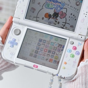 Puede incluir: Una consola de juegos portátil blanca con una funda protectora transparente que presenta pequeños iconos decorativos. La pantalla superior muestra una interfaz de juego de Hello Kitty, mientras que la pantalla táctil inferior muestra una cuadrícula.