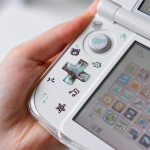 Op de afbeelding: Witte handheld gameconsole met decoratieve stickers, waaronder sterren, harten en een beer. De console heeft een D-pad, knoppen en een scherm met kleurrijke pictogrammen. Het apparaat wordt in een hand gehouden.