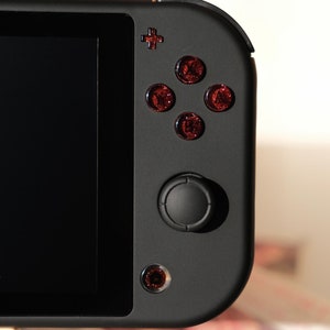 Pode incluir: Um console de jogos portátil Nintendo Switch preto com botões frontais vermelhos e um joystick preto.