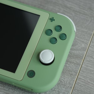 May include: A close-up shot of a handheld gaming console in a muted green color. The console features a black screen, green buttons, and a white joystick. The device is resting on a gray wooden surface.