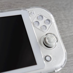 May include: A white handheld games console with a black screen, clear buttons, and a joystick. The device has a clear protective case with glitter. A pearl bead wrist strap is attached.