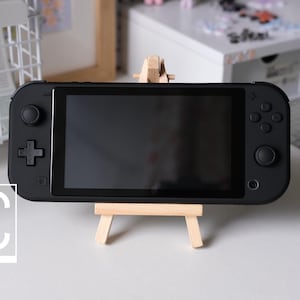 Może przedstawiać: Czarna przenośna konsola do gier Nintendo Switch z czarnym ekranem jest wyświetlana na małym drewnianym sztalugowym stojaku. Konsola ma czarno-białe logo z napisem "UC USMAN'S CONSOLES".