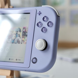 May include: A lavender handheld gaming console with a screen displaying a game interface. The console features a white joystick, translucent buttons, and a directional pad. The buttons are labeled with letters. The console is resting on a small wooden easel.