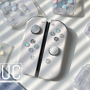 Può includere: Controller Joy-Con bianchi per Nintendo Switch con pulsanti personalizzati iridescenti e un pulsante a scacchi bianco e nero. I controller sono su una superficie bianca con altri piccoli contenitori di pulsanti.