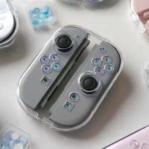 May include: A collection of gray, light blue, and pink game controllers with clear protective cases. The controllers have black joysticks and colorful button designs. Small clear containers with blue and clear decorative elements are also present.