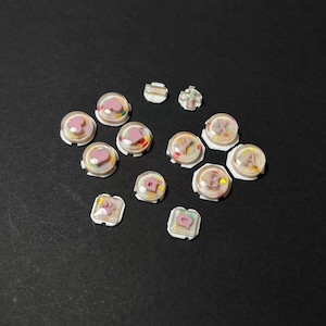 May include: A selection of clear, domed buttons with white bases. Some buttons feature pink hearts, letters, or floral designs. The buttons are scattered on a dark surface, offering a variety of decorative options.