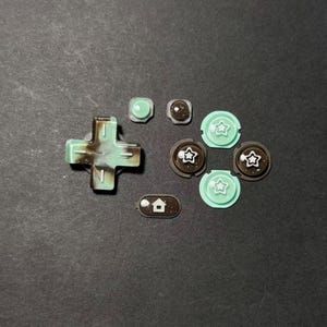 Peut inclure: Un ensemble de boutons de remplacement pour console de jeu de différentes formes et couleurs. L'ensemble comprend une croix directionnelle, des boutons ronds et des boutons avec des icônes d'étoile et de maison. Les boutons sont principalement vert menthe et marron.