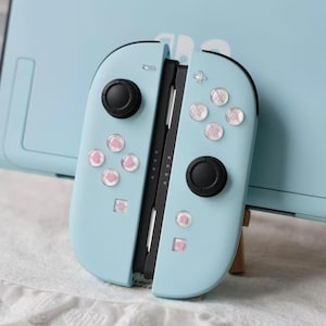 May include: A light blue video game controller with two black joysticks and buttons featuring small pink heart icons. The controller is positioned in front of a matching light blue console dock. The buttons have a clear, glossy finish over the pink hearts.