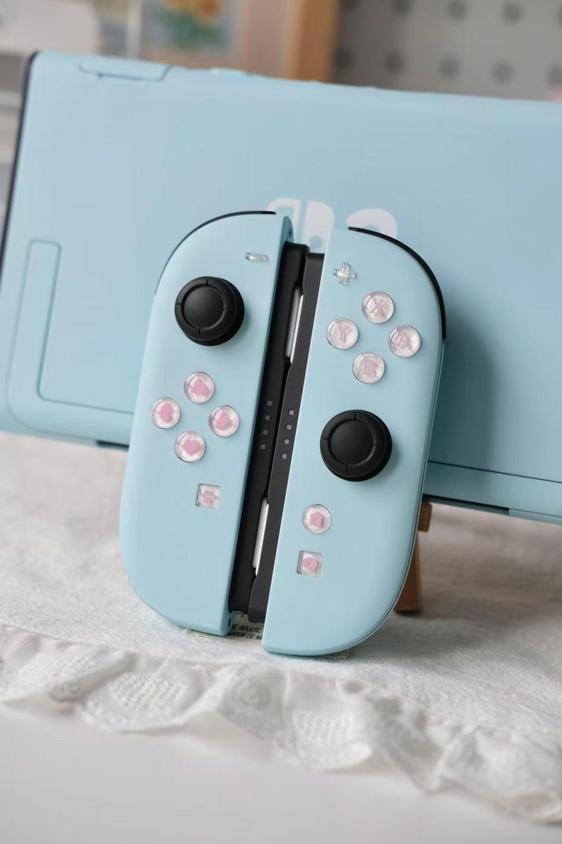 May include: A light blue video game controller with two black joysticks and buttons featuring small pink heart icons. The controller is positioned in front of a matching light blue console dock. The buttons have a clear, glossy finish over the pink hearts.