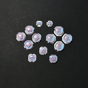 May include: A collection of iridescent game controller buttons in various shapes, including circles, squares, and a cross. The buttons feature a pink and glittery design with letters and symbols.