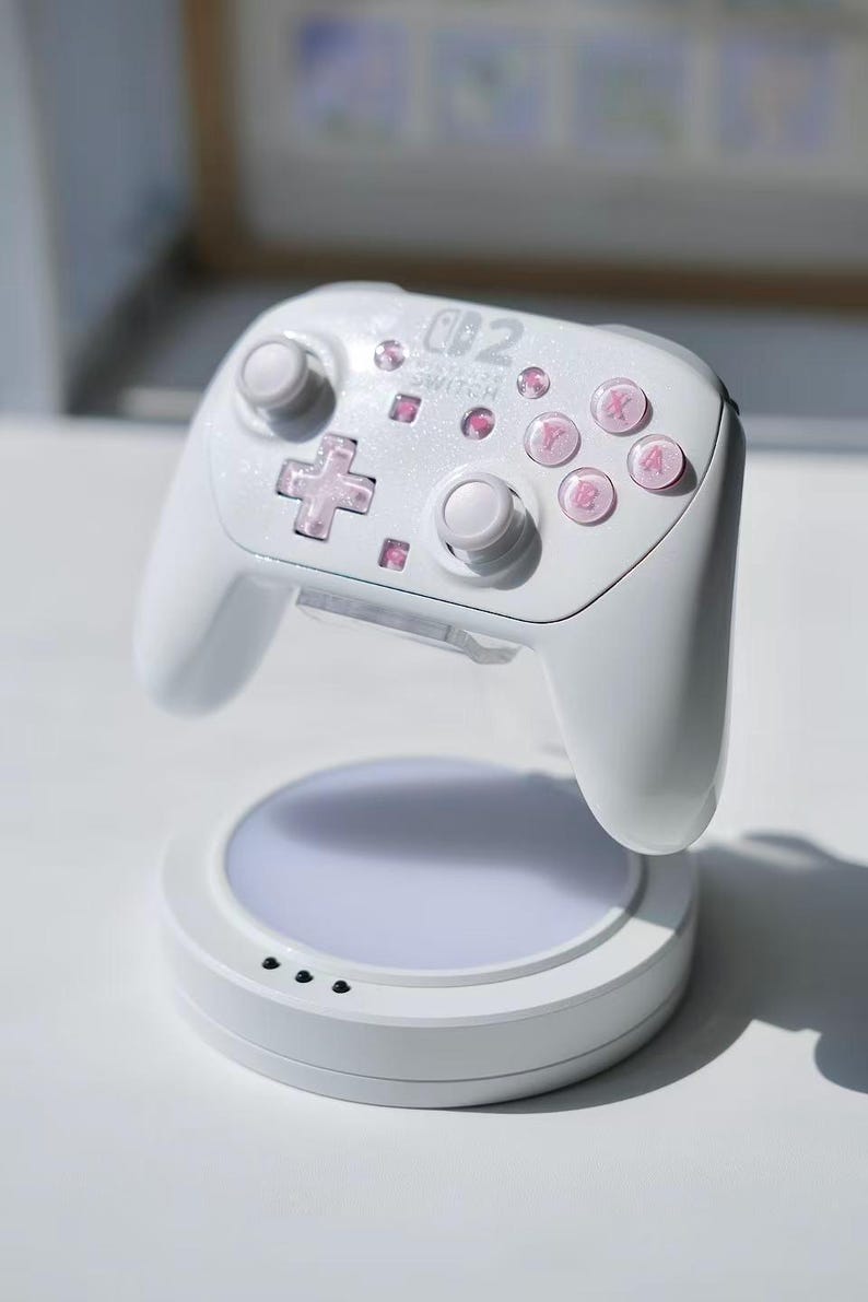 May include: A white wireless controller for a gaming console featuring a glittery finish and translucent pink buttons. The controller is mounted on a circular white charging dock with a flat, illuminated base and three small black indicator lights.