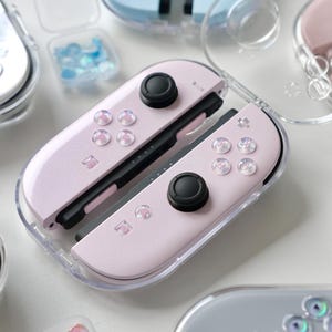 Könnte beinhalten: Nahaufnahme mehrerer pastellfarbener Nintendo Switch Joy-Cons in transparenten Schutzhüllen. Die Joy-Cons sind hellrosa, blau und grau, mit schwarzen Steuerknüppeln und dekorativen Knöpfen. Kleine Behälter mit schillernden Verzierungen sind ebenfalls sichtbar.