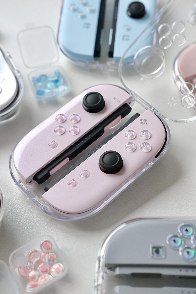 May include: Close-up of pastel-coloured Nintendo Switch Joy-Cons in clear protective cases. The Joy-Cons are light pink, blue, and grey, with black control sticks and decorative buttons. Small containers of iridescent embellishments are also visible.