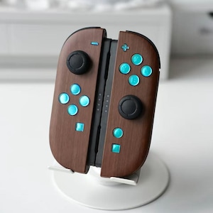 May include: A pair of video game controllers with a dark brown wood grain finish. The controllers feature bright cyan buttons, black analog sticks, and a central black connector rail. The device is mounted on a white circular stand.