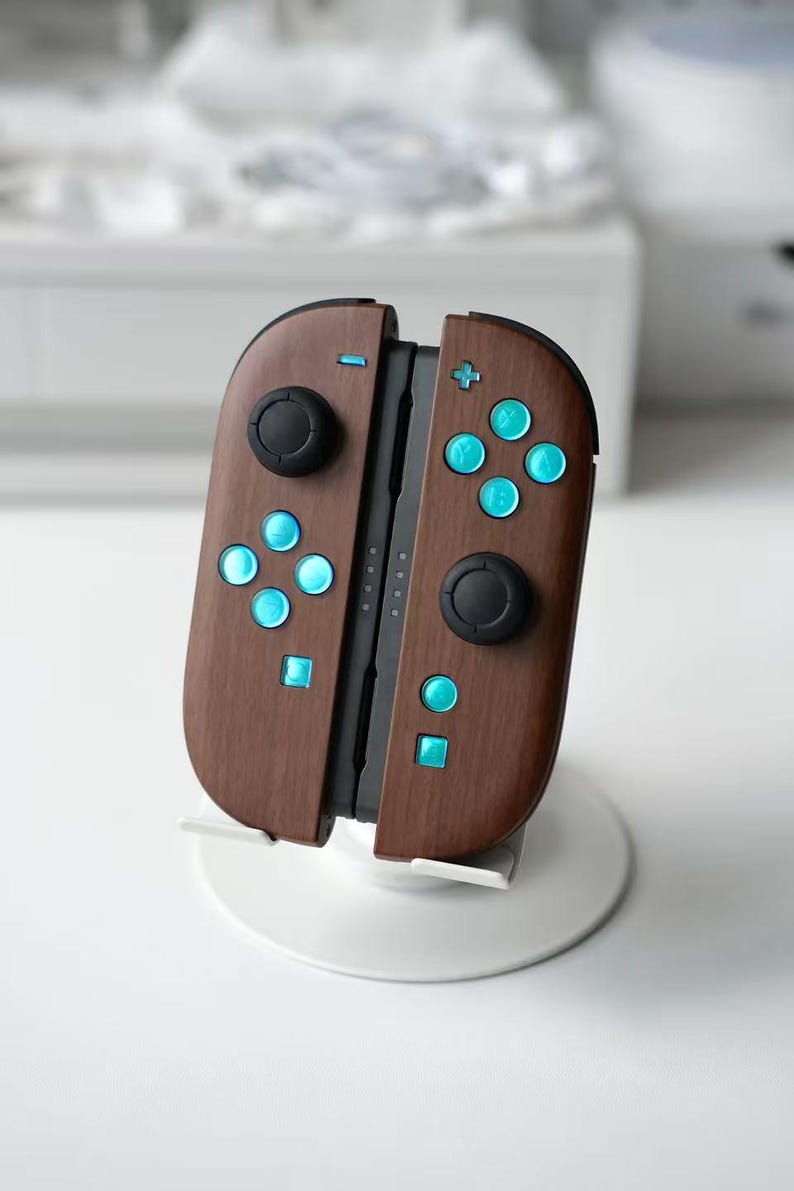 May include: A pair of video game controllers with a dark brown wood grain finish. The controllers feature bright cyan buttons, black analog sticks, and a central black connector rail. The device is mounted on a white circular stand.