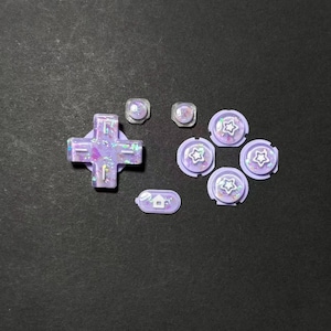 May include: A collection of custom controller buttons in shades of purple, with iridescent glitter accents. Includes a directional pad, several round buttons with star designs, and smaller buttons with a house icon.