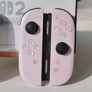 Może przedstawiać: Para różowych kontrolerów Joy-Con do Nintendo Switch z czarnymi joystickami i przyciskami. Kontrolery mają błyszczące wykończenie i są umieszczone na białym tle. Logo Nintendo Switch jest widoczne w tle.