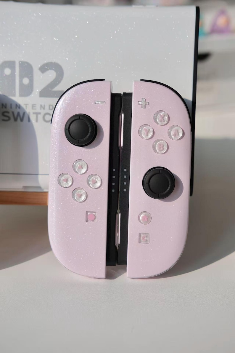 May include: Pink Nintendo Switch Joy-Con controllers with black joysticks and buttons. The controllers have a glittery finish. The Nintendo Switch logo is visible in the background. The controllers are set against a white background.