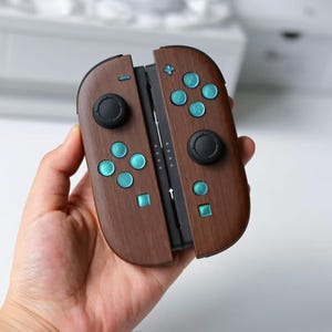 May include: A pair of game controllers featuring a dark brown wood grain finish. The device includes two black circular joysticks, eight metallic teal buttons with letter markings, and two small square buttons. The controllers are joined together.