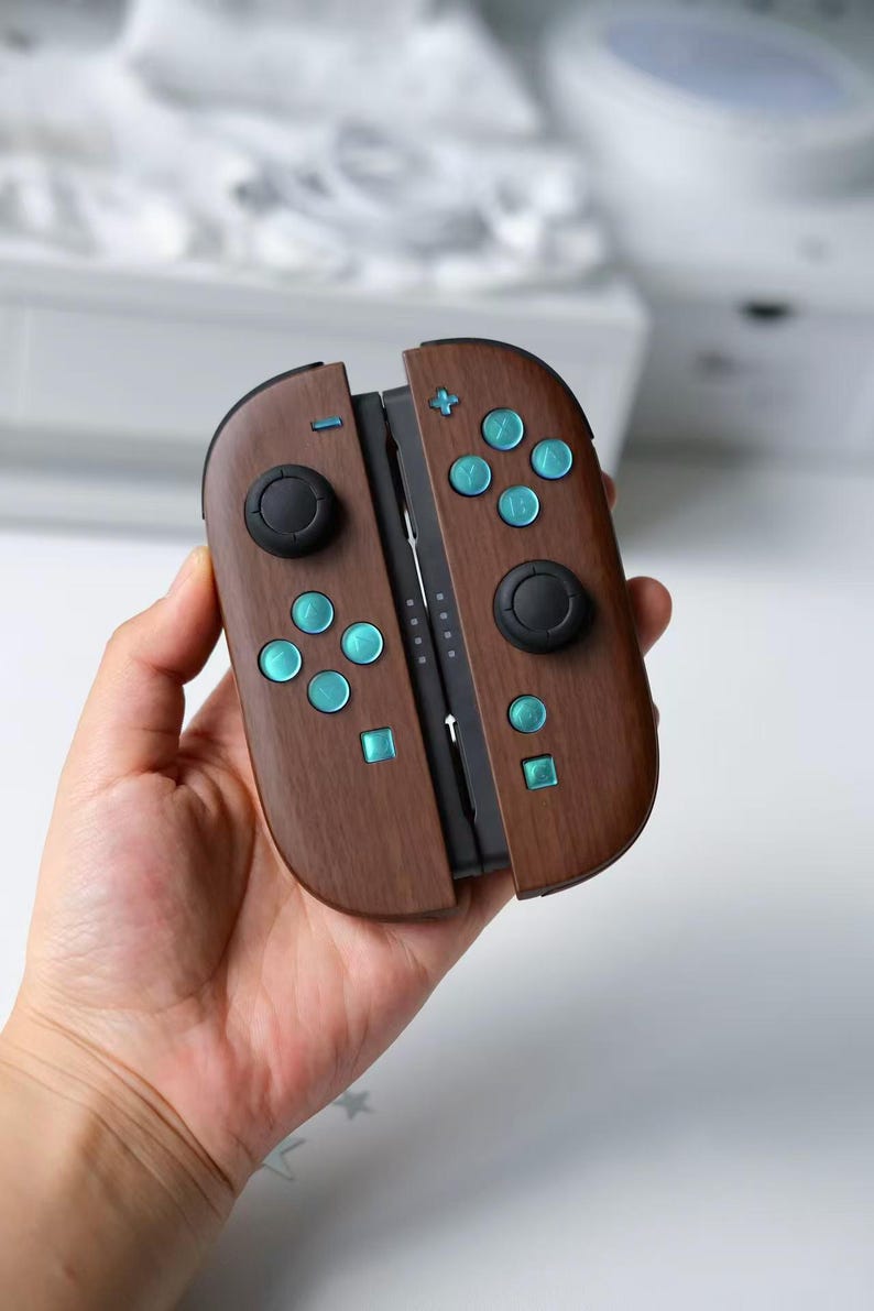 May include: A pair of game controllers featuring a dark brown wood grain finish. The device includes two black circular joysticks, eight metallic teal buttons with letter markings, and two small square buttons. The controllers are joined together.