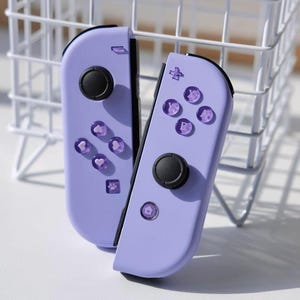 以下が含まれることがあります： 黒いコントロールパッドと半透明の紫色のボタンが付いた、2つのラベンダー色のゲームコントローラー。 コントローラーは白いワイヤーバスケットに寄りかかっています。 ボタンには文字と記号のデザインがあります。