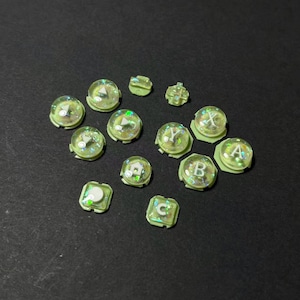 May include: A collection of translucent green and clear buttons with iridescent glitter. Some buttons have letters A, B, C, X, and Y. The buttons are of various shapes and sizes, including round and square, and are set against a dark background.