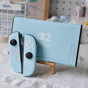 May include: A light blue Nintendo Switch console and two matching Joy-Con controllers. The console features the white Nintendo Switch logo and the number 2 on the back. The controllers have light blue shells with black joysticks and buttons.