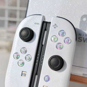 May include: White Nintendo Switch Joy-Con controllers with iridescent buttons and black joysticks. The controllers have a glittery finish. The Nintendo Switch logo is visible in the background.