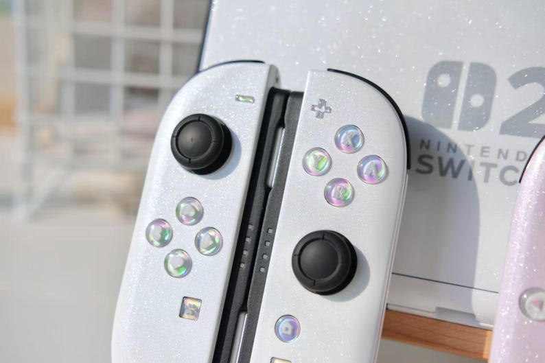 May include: White Nintendo Switch Joy-Con controllers with iridescent buttons and black joysticks. The controllers have a glittery finish. The Nintendo Switch logo is visible in the background.