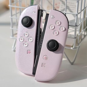 May include: A pair of pale pink Nintendo Switch Joy-Con controllers. The controllers have a glittery finish and feature black control sticks and clear buttons with symbols. The controllers are designed for gaming.