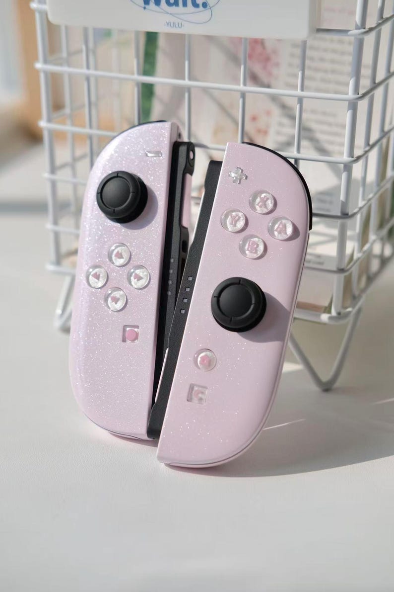 May include: A pair of pale pink Nintendo Switch Joy-Con controllers. The controllers have a glittery finish and feature black control sticks and clear buttons with symbols. The controllers are designed for gaming.