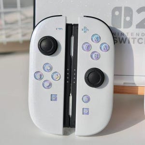 Puede incluir: Mandos Joy-Con blancos de Nintendo Switch con botones negros y detalles iridiscentes. Los mandos tienen un acabado brillante y presentan la disposición estándar de los botones. El logotipo de Nintendo Switch es visible en el fondo.