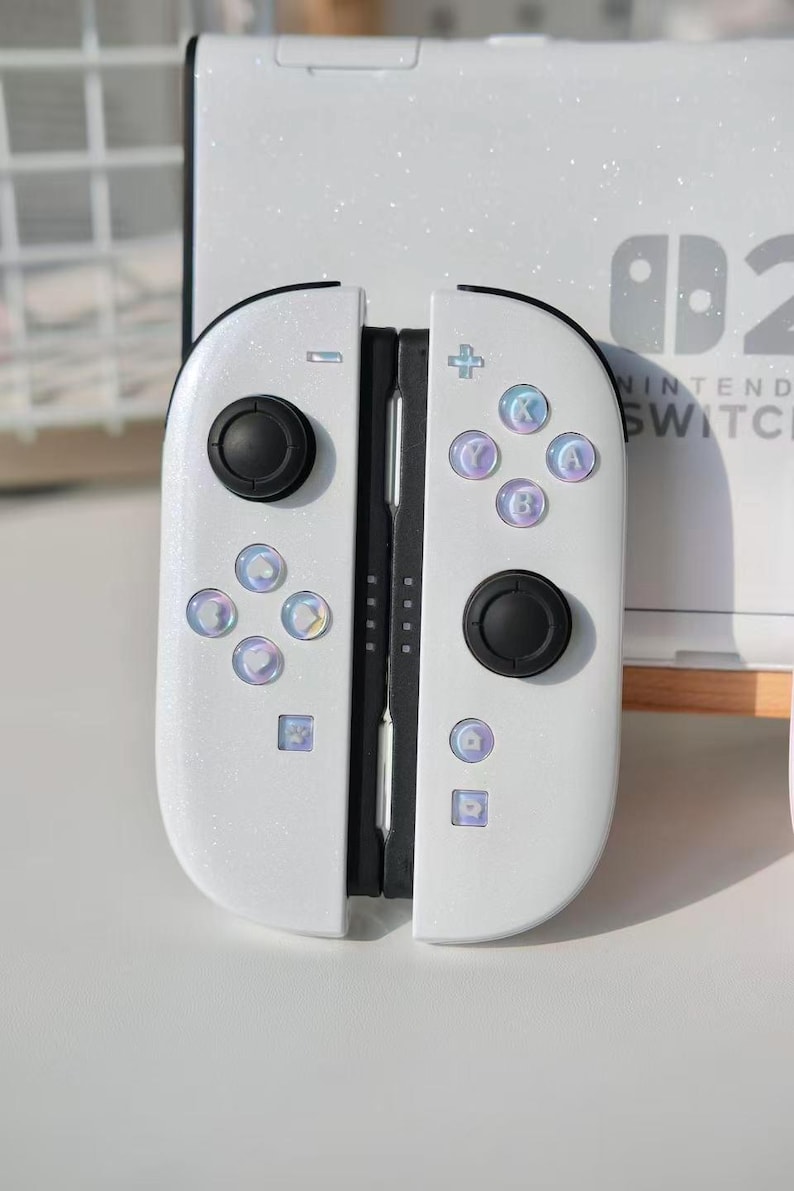 May include: White Nintendo Switch Joy-Con controllers with black buttons and iridescent accents. The controllers have a glittery finish and feature the standard button layout. The Nintendo Switch console is visible in the background.