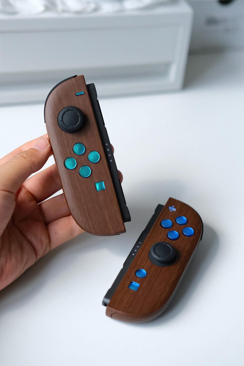 May include: Two game controller attachments featuring a dark brown wood grain finish. Each unit includes a black circular joystick, a set of bright blue circular buttons, and a single blue square button. The controllers have a sleek, curved design.
