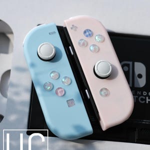 May include: A pair of pastel blue and pink game controllers with white buttons and iridescent gems. The controllers are for a gaming console, with the text "USMAN'S CONSOLES" visible.