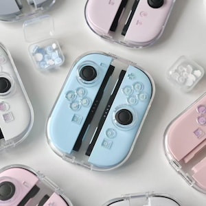 May include: A selection of pastel-coloured Nintendo Switch Joy-Con controllers in clear protective cases. The controllers are in shades of pink, blue, and white, with black joysticks and buttons. Small containers of matching coloured thumb grips are also visible.