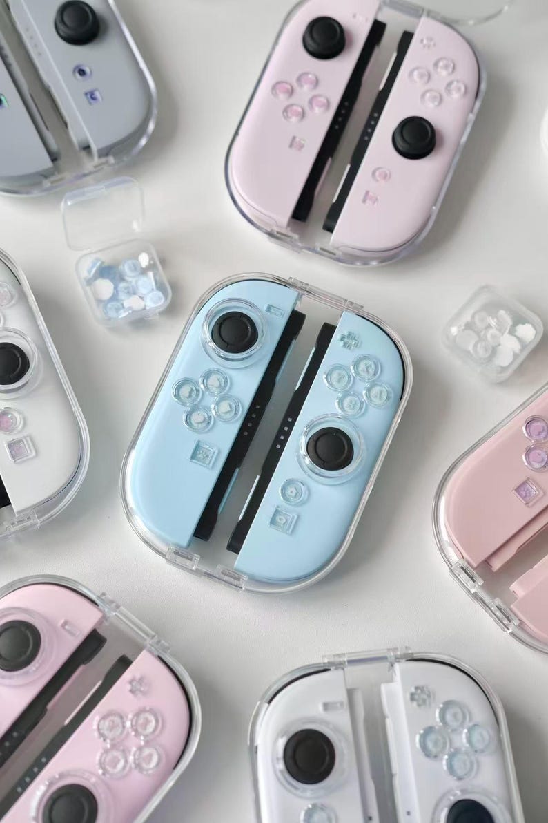 May include: A selection of pastel-coloured Nintendo Switch Joy-Con controllers in clear protective cases. The controllers are in shades of pink, blue, and white, with black joysticks and buttons. Small containers of matching coloured thumb grips are also visible.